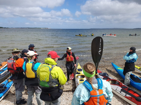 A group of kayakers receives instruction on a beach near Roslev, Denmark, with kayaks and sea view.