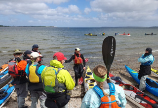A group of kayakers receives instruction on a beach near Roslev, Denmark, with kayaks and sea view.