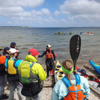 A group of kayakers receives instruction on a beach near Roslev, Denmark, with kayaks and sea view.