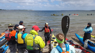 A group of kayakers receives instruction on a beach near Roslev, Denmark, with kayaks and sea view.