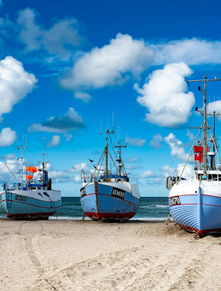 Fishing boats pulled onto the sandy beach near Aars in North Jutland, Denmark, under a bright blue sky.
