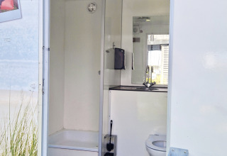 Glampingbadeværelse med bruser, toilet og håndvask, set gennem en åben dør i dagslys.