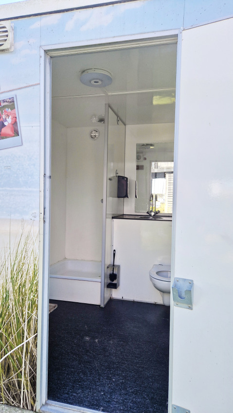 Glampingbadkamer met douche, toilet en wastafel, gezien door een open deur bij daglicht.