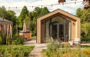 Modernes Tiny House aus Holz mit Terrasse, großen Fenstern und Außen-Whirlpool in grüner Umgebung.