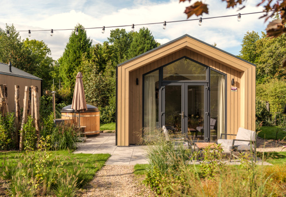 Tiny house moderne en bois avec grandes fenêtres, terrasse, coin salon extérieur et bain à remous au jardin.
