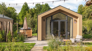 Modernes Tiny House aus Holz mit Terrasse, großen Fenstern und Außen-Whirlpool in grüner Umgebung.