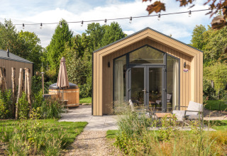 Moderne tiny house i træ med store vinduer, terrasse og udendørs hot tub omgivet af grøn have.