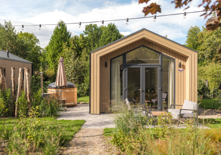 Moderne tiny house van hout met grote ramen, buitenterras, zitgedeelte en een hot tub in groene tuin.