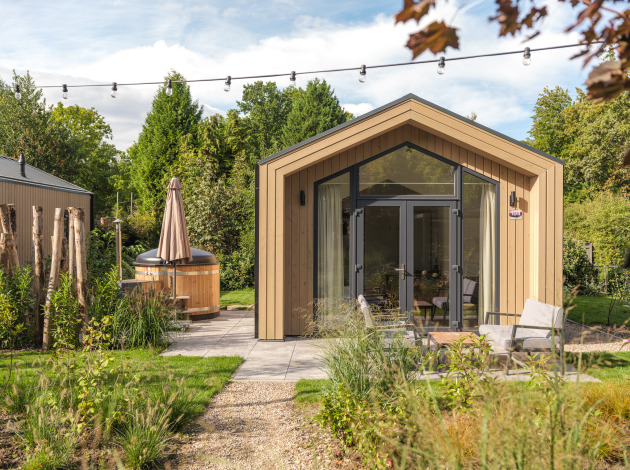Moderne tiny house van hout met grote ramen, buitenterras, zitgedeelte en een hot tub in groene tuin.