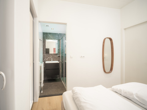 Bedroom with modern bathroom, wall mirror, and white bedding in Lodge Essenburgh at Veluwepark de Bosgraaf.