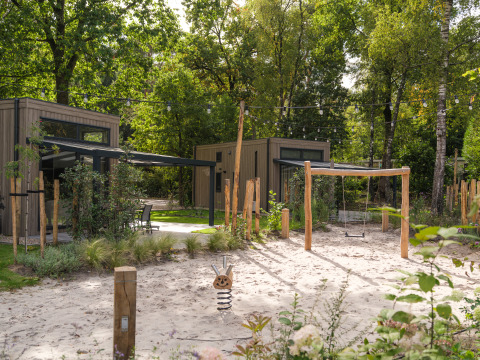 Buitenspeeltuin met zand, schommel en houten huisjes aan een lodge, omgeven door groene bomen.