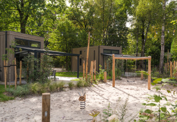 Udendørs legeplads med sand, gynger og små træhytter ved en lodge omgivet af grønne træer.