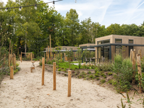 Moderne Lodge mit Holzfassade, Außensitzbereich, Sandweg und Spielplatz, umgeben von Bäumen.