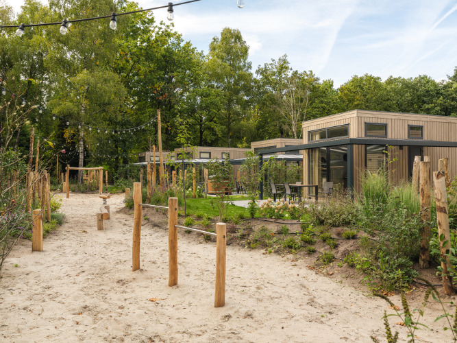 Moderne Lodge mit Holzfassade, Außensitzbereich, Sandweg und Spielplatz, umgeben von Bäumen.