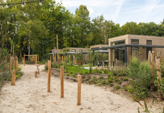 Moderne Lodge mit Holzfassade, Außensitzbereich, Sandweg und Spielplatz, umgeben von Bäumen.