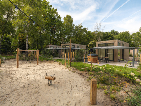 Moderne lodge met privéterras, jacuzzi en zand-speelplaats met schommel, omringd door groene bomen.