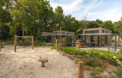 Modern lodge with private patio, hot tub, and sandy playground area with swings, surrounded by trees.