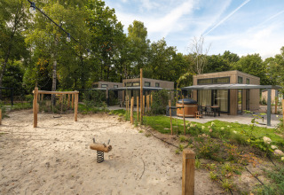 Moderne lodge met privéterras, jacuzzi en zand-speelplaats met schommel, omringd door groene bomen.
