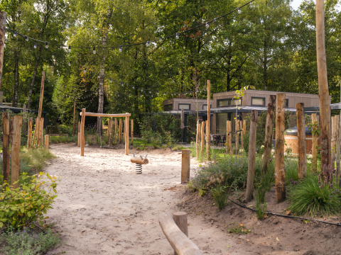 Zandige speeltuin met houten toestellen en lichtslingers aan een lodge tussen bomen en natuur.