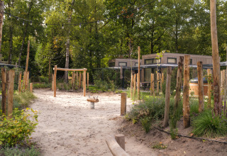 Zandige speeltuin met houten toestellen en lichtslingers aan een lodge tussen bomen en natuur.