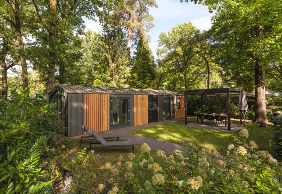Moderne Cozy Chalet lodge blandt træerne med liggestol og overdækket spiseplads i grønne omgivelser.