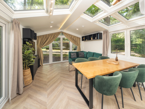 Lyst og moderne chalet-interiør med store vinduer, spisebord og grønne stole i Veluwepark de Bosgraaf, Nederlandene.