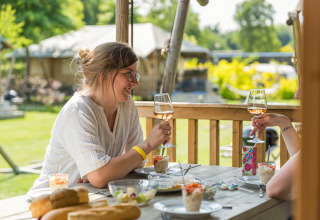 Zwei Frauen genießen Wein und Essen auf der Terrasse eines Safarizeltes im Vakantiepark Ackersate, Niederlande.