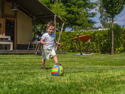 Niño pequeño juega con una pelota colorida en el césped junto a una tienda safari en Vakantiepark Ackersate.