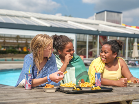Tres niños con toallas disfrutan de papas fritas y refrescos junto a la piscina en un parque vacacional.