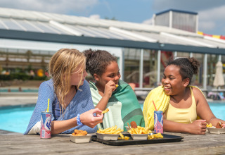 Tres niños con toallas disfrutan de papas fritas y refrescos junto a la piscina en un parque vacacional.