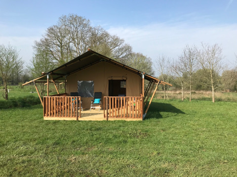 Safari-Zelt mit Holzveranda auf einer grünen Wiese, von Bäumen umgeben, unter blauem Himmel.