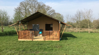 Tenda safari con veranda in legno su prato verde, circondata da alberi e cielo azzurro sereno.