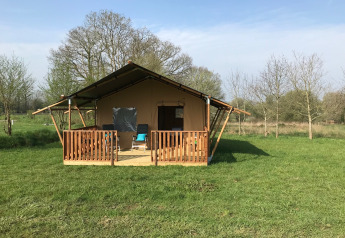 Tenda safari con veranda in legno su prato verde, circondata da alberi e cielo azzurro sereno.