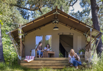 Fire kvinder slapper af og hygger sig foran et safaritelt i skoven med træer og solskin omkring dem.