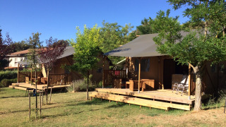 Safari lodge tents with private sanitary facilities, wooden decks, and chairs set in a green, leafy campground.