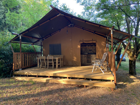 Tenda safari con servizi igienici, mobili in legno sulla veranda e circondata da natura verde e alberi.