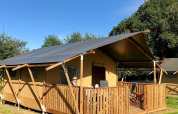 Tenda safari al Camping O2 in Francia con veranda in legno, sedie e tanto verde attorno sotto cielo sereno.