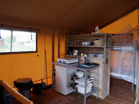 Interior de una tienda safari con cocina pequeña, estantería de madera, nevera y litera al fondo.