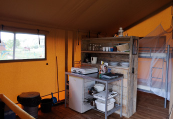 Interior de una tienda safari con cocina pequeña, estantería de madera, nevera y litera al fondo.