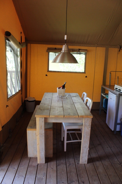 A wooden table with chairs and bench under a metal lamp inside a safari tent with wooden flooring.