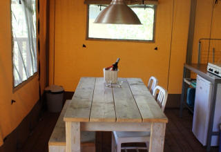 A wooden table with chairs and bench under a metal lamp inside a safari tent with wooden flooring.