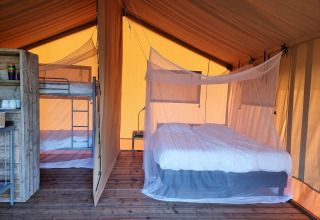 Interieur van een safaritent met houten vloer, tweepersoonsbed met klamboe en stapelbed, gescheiden door doek.