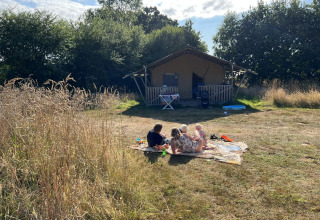 En familie nyder en solrig dag udenfor et safaritelt på Camping O2 i Frankrig, omgivet af natur.