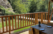 View from a safari tent terrace with wooden furniture, green hills, and blue sky at a French campsite.