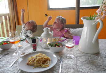 Due bambini mangiano pasta a tavola in una sistemazione glamping con insalata, bevande e atmosfera accogliente.