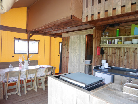 Interieur van een safari lodgetent met houten meubels, eettafel, keuken en eigen sanitair.