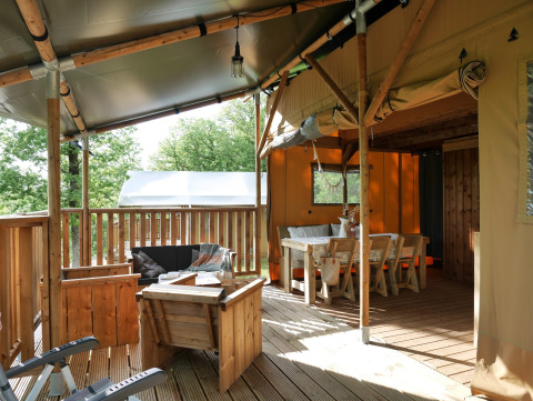 Überdachte Terrasse eines Glamping-Zeltes mit Holzmöbeln, Lounge und Essbereich inmitten grüner Natur.