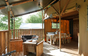 Overdekt terras bij een glampingverblijf met houten meubilair, zithoek en eettafel midden in de natuur.