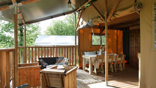 Covered deck area at a glamping site, featuring wooden furniture, a lounge, and a dining table in nature.