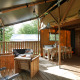 Covered deck area at a glamping site, featuring wooden furniture, a lounge, and a dining table in nature.